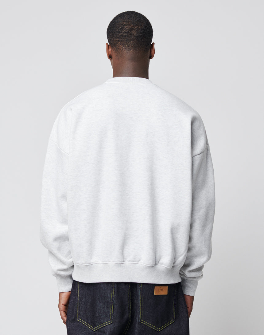 A person stands facing away from the camera, wearing the LIVE FAST DIE YOUNG Live Fast Die Young Chest Sweater in light grey with visible stitching and a subtle vintage effect, paired with dark blue jeans against a plain light grey background.