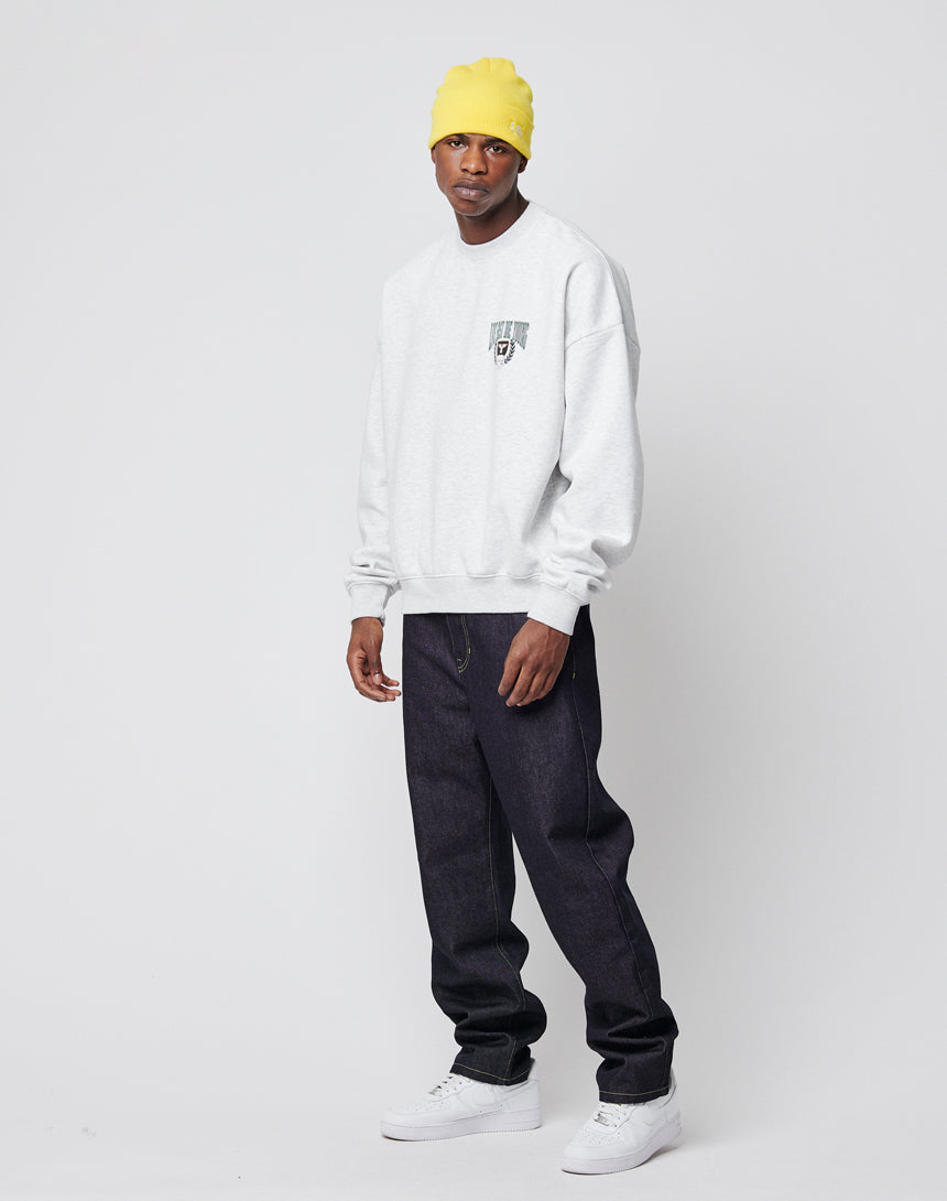 A man stands against a plain white background wearing the LIVE FAST DIE YOUNG Live Fast Die Young Chest Sweater, a light gray sweater with a vintage effect and small chest graphic, paired with a yellow beanie, dark jeans, and white sneakers.