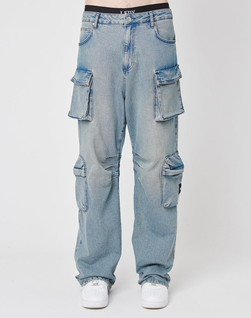 A person sports LIVE FAST DIE YOUNG’s Wide Denim Cargo in light blue with side pockets and a faded vintage effect, styled with white sneakers. The relaxed, oversized fit jeans are shown from the waist down against a plain background.