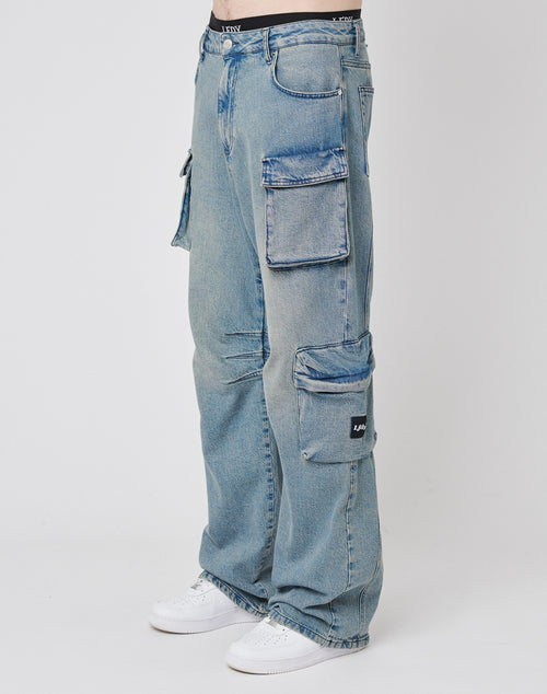 A person wears LIVE FAST DIE YOUNG’s Wide Denim Cargo jeans—loose-fitting, light blue denim with large side pockets—paired with white sneakers for a relaxed streetwear look.