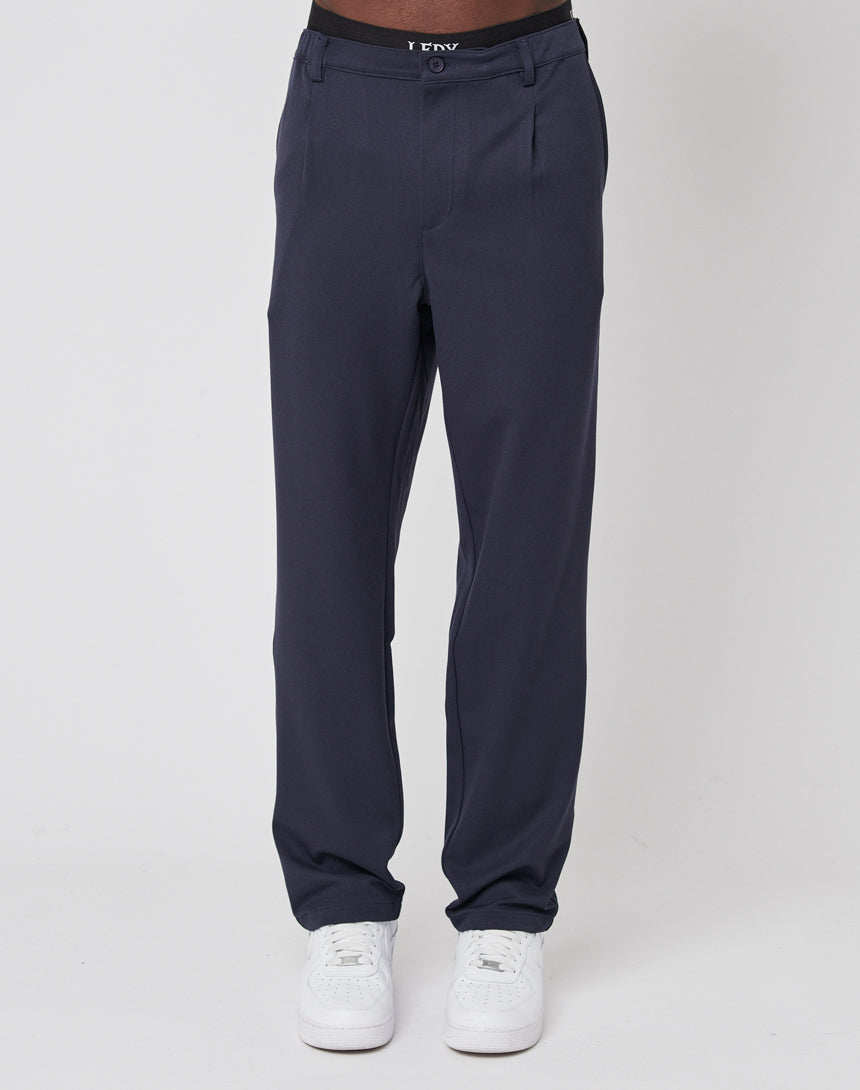 A person wears LIVE FAST DIE YOUNG Regular Casual Pants with an elastischer Bund and white sneakers, standing against a plain white background. Only the lower half of the body is visible.
