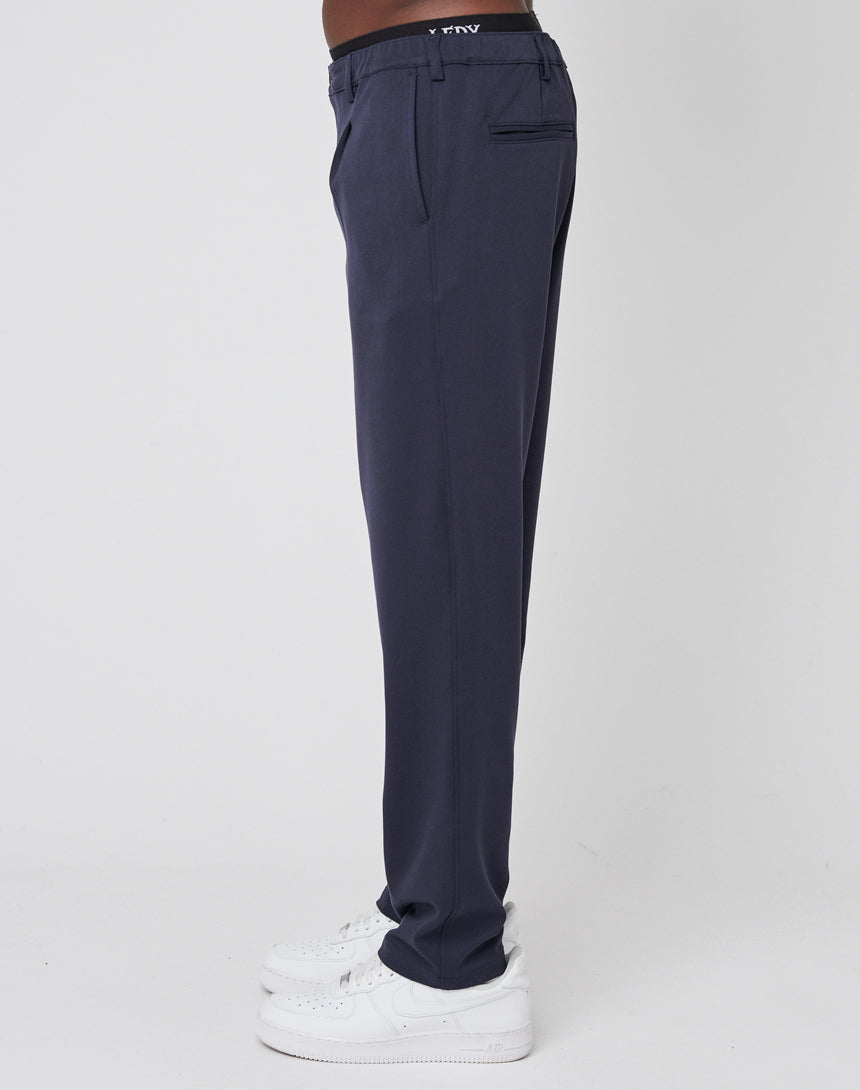 A person stands sideways in Regular Casual Pants (navy, regular fit) from LIVE FAST DIE YOUNG, paired with white sneakers and a black elastic waistband featuring the brand name. The background is plain white.