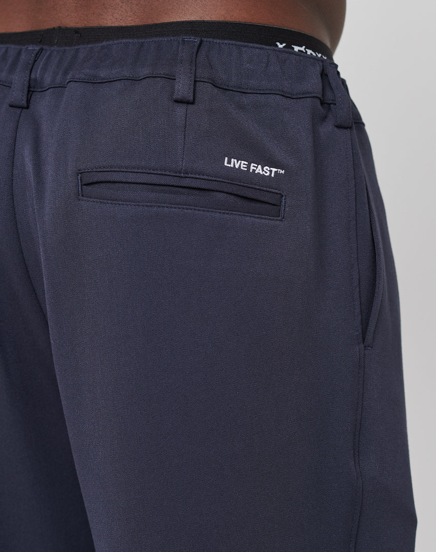 Close-up of the back and side of Regular Casual Pants by LIVE FAST DIE YOUNG in navy blue, featuring a welt back pocket with “LIVE FAST™” embroidered above it in white and an elastic waistband, worn by a person with medium brown skin.
