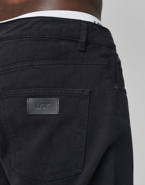 Close-up of a person wearing Regular Denim by LIVE FAST DIE YOUNG. Features a black LDY patch, tapered leg, regular fit, and back pocket detail. White underwear waistband is visible. Made from 100% Baumwolle for comfort.