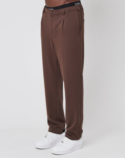 A person wearing LIVE FAST DIE YOUNG Regular Casual Pants with pleats, an elastic waist, a black LFDY waistband, and white sneakers stands against a plain white background.