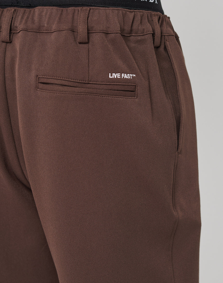 Close-up of brown Regular Casual Pants by LIVE FAST DIE YOUNG featuring a side pocket, back welt pocket, and LIVE FAST™ printed in white above the back pocket.