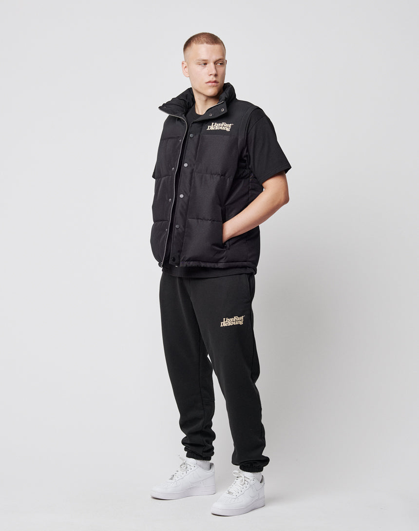 A man stands against a plain background wearing the LIVE FAST DIE YOUNG Basic Vest over a black T-shirt, Regular Fit jogger pants, and white sneakers. His hands are in his vest pockets as he looks slightly to the side.