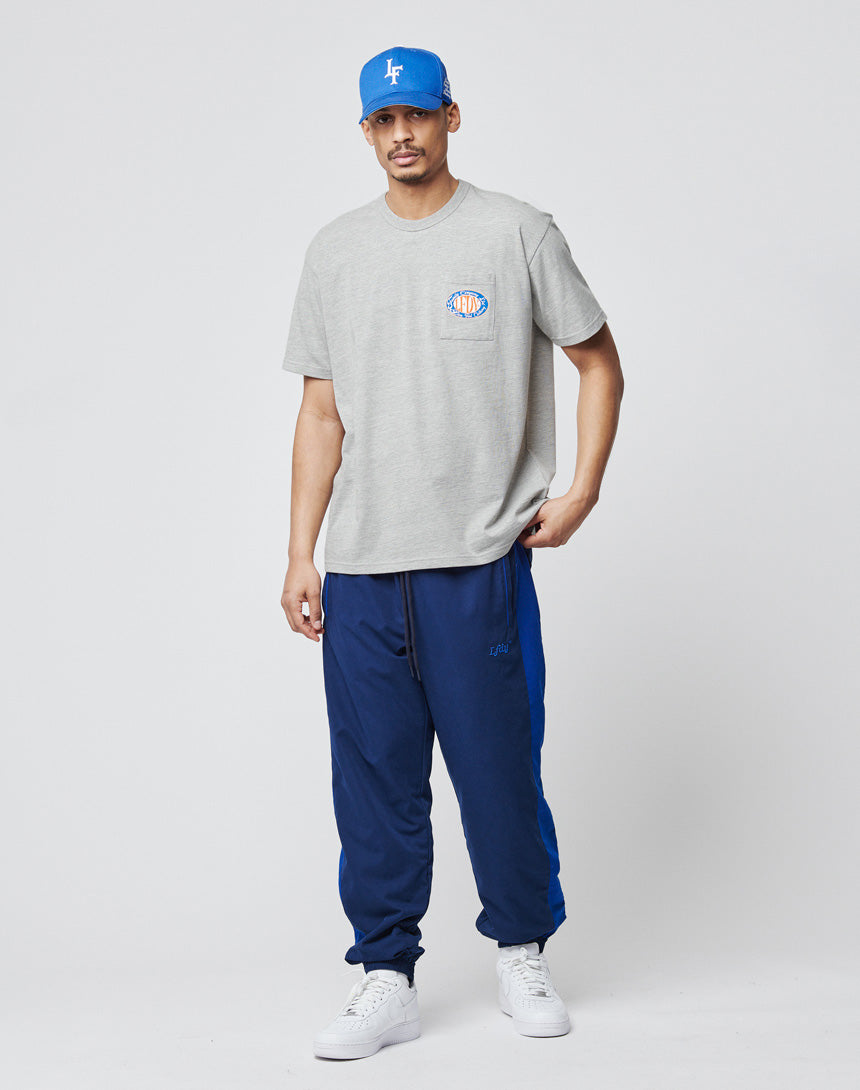 A man stands against a plain background wearing a blue cap, the LIVE FAST DIE YOUNG Family Pocket Tee in light grey with a small chest logo, blue track pants with side stripes, and white sneakers. His left hand is in his pocket.