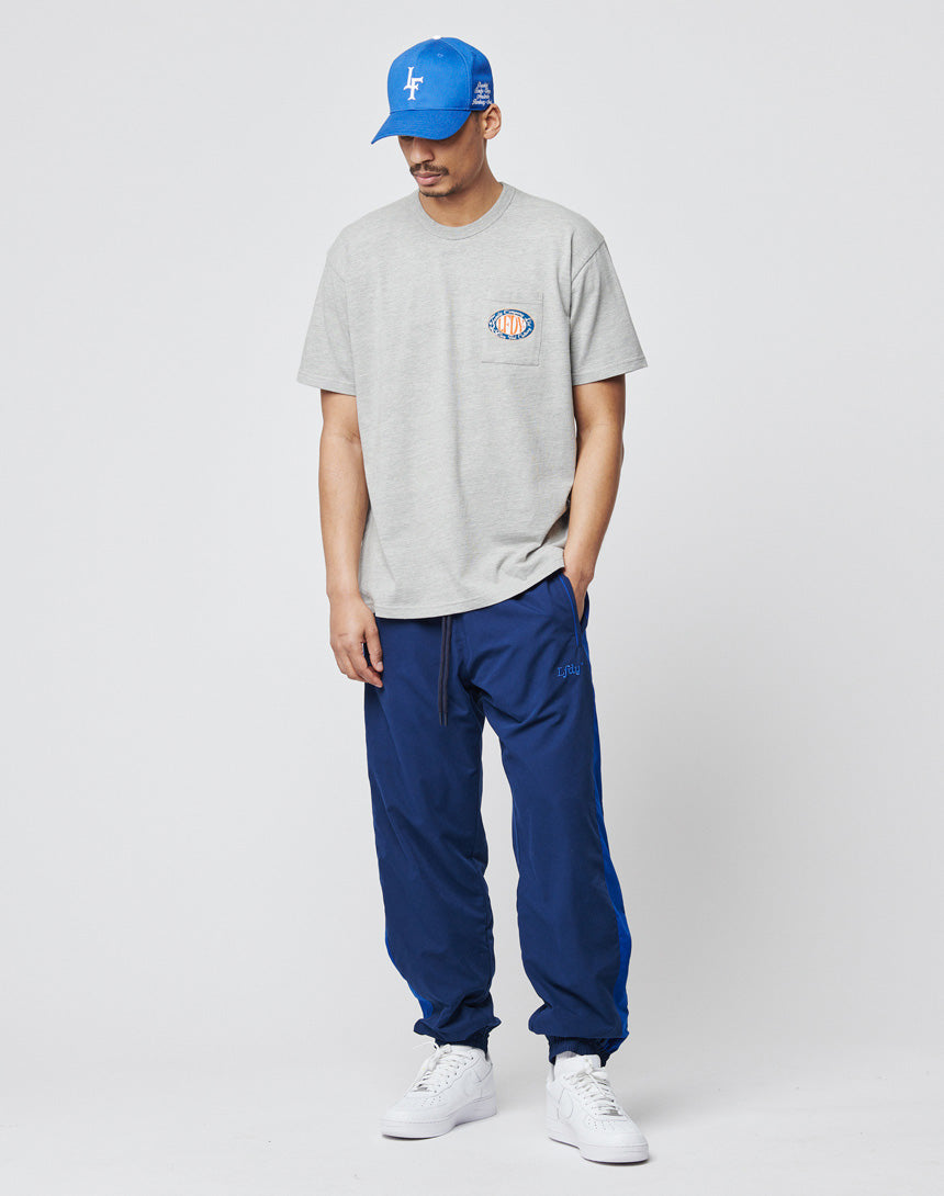 A man in a blue cap, light gray polyester t-shirt with a small chest graphic, LFDY Trackpants by LIVE FAST DIE YOUNG, and white sneakers stands against a plain white background.