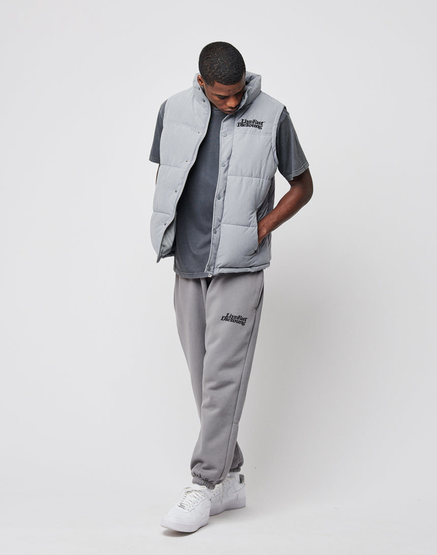A man wearing a LIVE FAST DIE YOUNG Basic Vest in light gray over a dark gray t-shirt, paired with gray sweatpants and white sneakers, stands against a white background looking down, with his hands in his vest pockets.