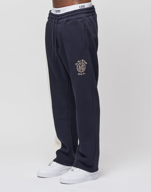 A person wears LIVE FAST DIE YOUNGs LFDY Split Jogger—navy blue with a beige side stripe, embroidered crest and text on the thigh, wide fit, and elastic waist—paired with white sneakers against a plain white background.