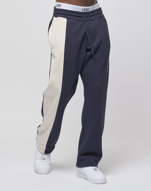 A person wears the LFDY Split Jogger by LIVE FAST DIE YOUNG, featuring navy blue fabric with a wide beige stripe down one leg, a Wide Fit, elastic waist with LFDY print, paired with white sneakers against a plain white background.