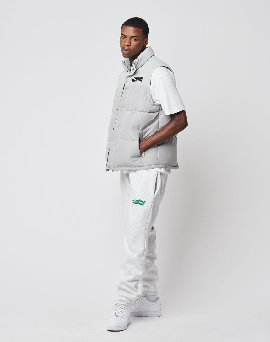 A man stands against a white background wearing the LIVE FAST DIE YOUNG Basic Vest in light gray polyester, paired with a white T-shirt, gray sweatpants, and white sneakers. He has his left hand in his pocket and looks slightly to the left. Regular fit.