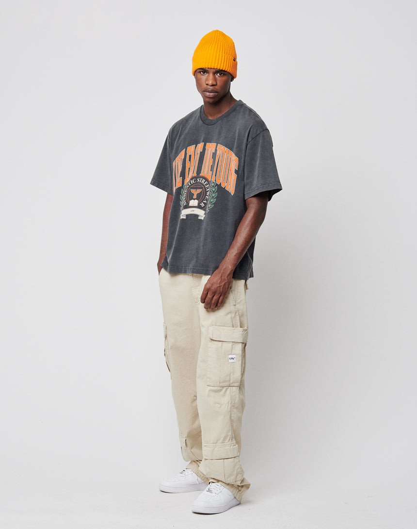 A man wearing the Live Fast Die Young Tee by LIVE FAST DIE YOUNG, a yellow beanie, beige cargo pants, and white sneakers stands against a plain white background.