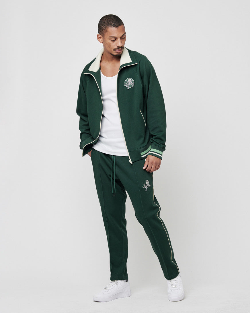 A man poses against a white background in a LIVE FAST DIE YOUNG Track Suit Jacket, Regular Fit, dark green with light green accents and a hoher Kragen. He pairs it with a white tank top and sneakers, looking down to his left with one hand in his pocket.
