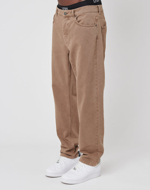 A person wears LIVE FAST DIE YOUNGs Regular Denim—loose-fitting brown jeans with a tapered leg, black waistband with white text, and regular fit made from 100% Baumwolle—plus white sneakers, standing against a plain white background.