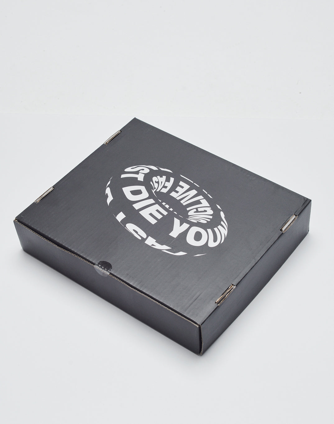 A closed black cardboard box featuring a warped white circular LIVE FAST DIE YOUNG logo on the lid, containing the Live Fast Swim Ring, is placed on a plain white surface.