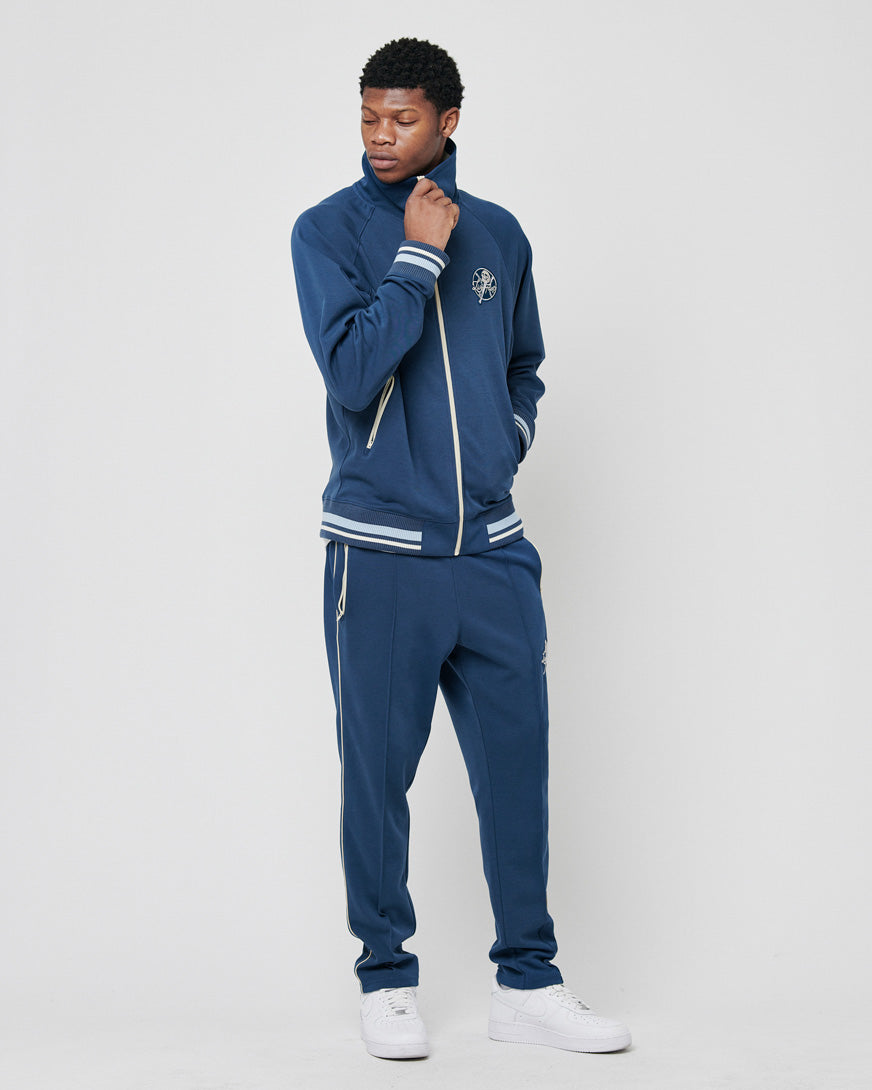 A man poses against a plain background wearing a LIVE FAST DIE YOUNG Track Suit Jacket in navy, with matching pants featuring white stripes on the cuffs and collar. The set is made from soft polyester-cotton and paired with white sneakers. One hand rests near his chin.