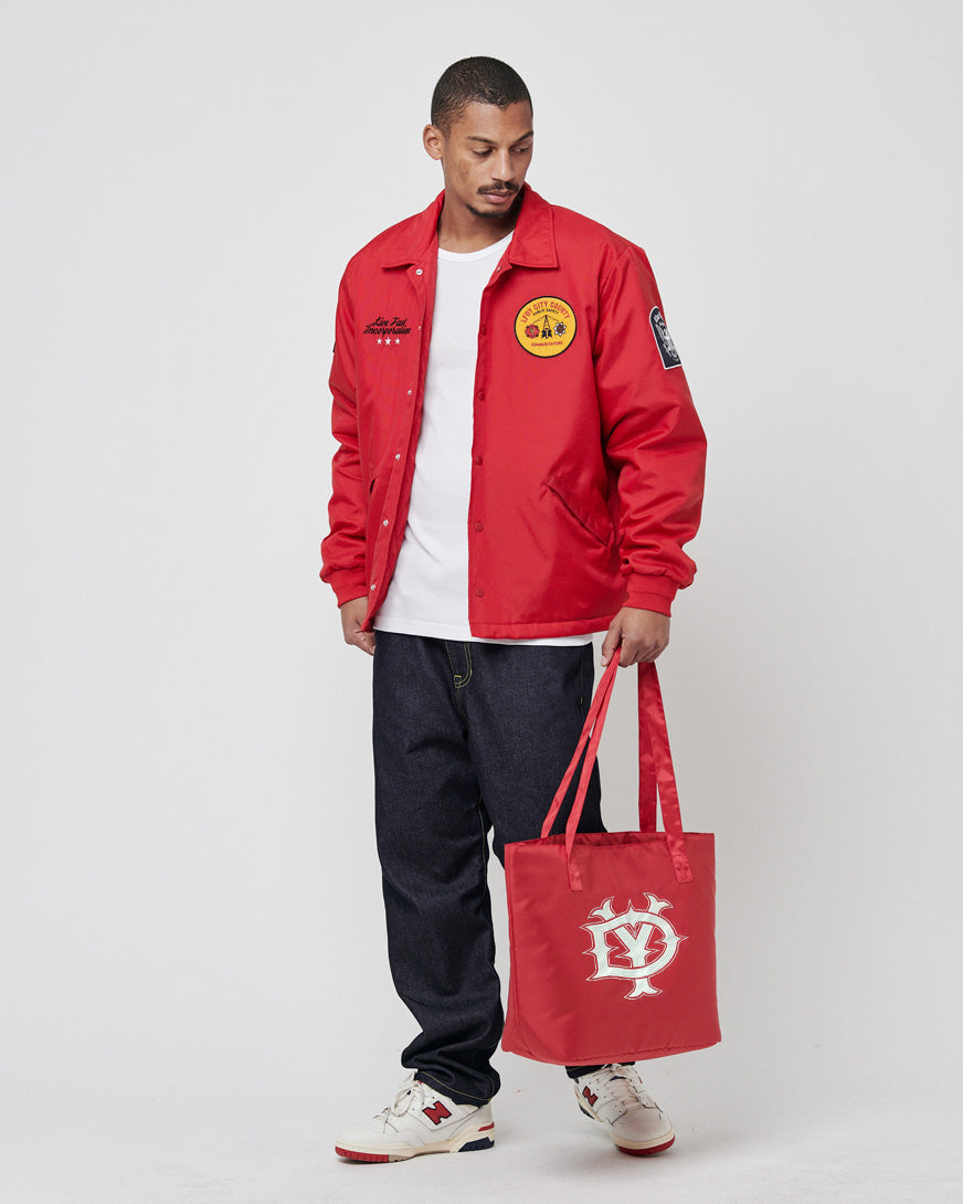 A man stands against a plain background wearing a leicht wattierte LFDY Coach Jacket by LIVE FAST DIE YOUNG, a white T-shirt, dark jeans, and white sneakers. He holds a red tote bag with a white logo in his left hand, looking slightly downward.
