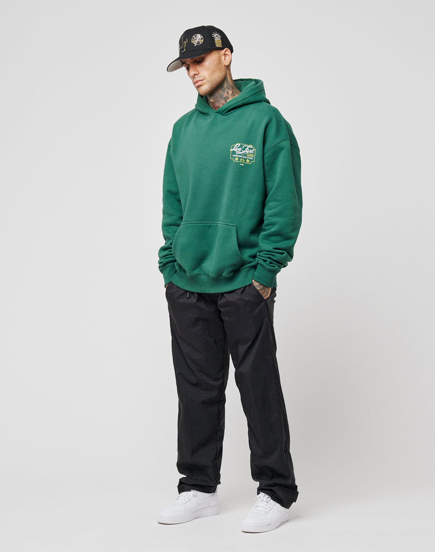 A man wearing the LIVE FAST DIE YOUNG Live Fast Greetings Hooded in dark green, with black pants, white sneakers, and a black cap stands with his hands in his pockets against a plain white background.