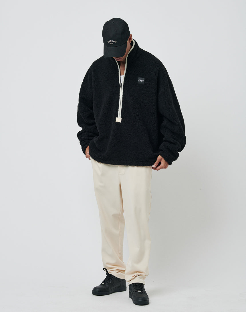 A person wearing the LFDY Teddy Half Zip black fleece pullover, cream pants, black shoes, and a black baseball cap stands against a white background, looking down with hands in their pockets.