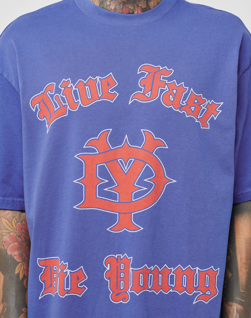 A person wears the LIVE FAST DIE YOUNG DY Tee (blue, 100% Baumwolle, Regular Fit) featuring red and white gothic Live Fast Die Young text and a bold DY symbol. Visible neck and arm tattoos highlight its Vintage-Effekt style.