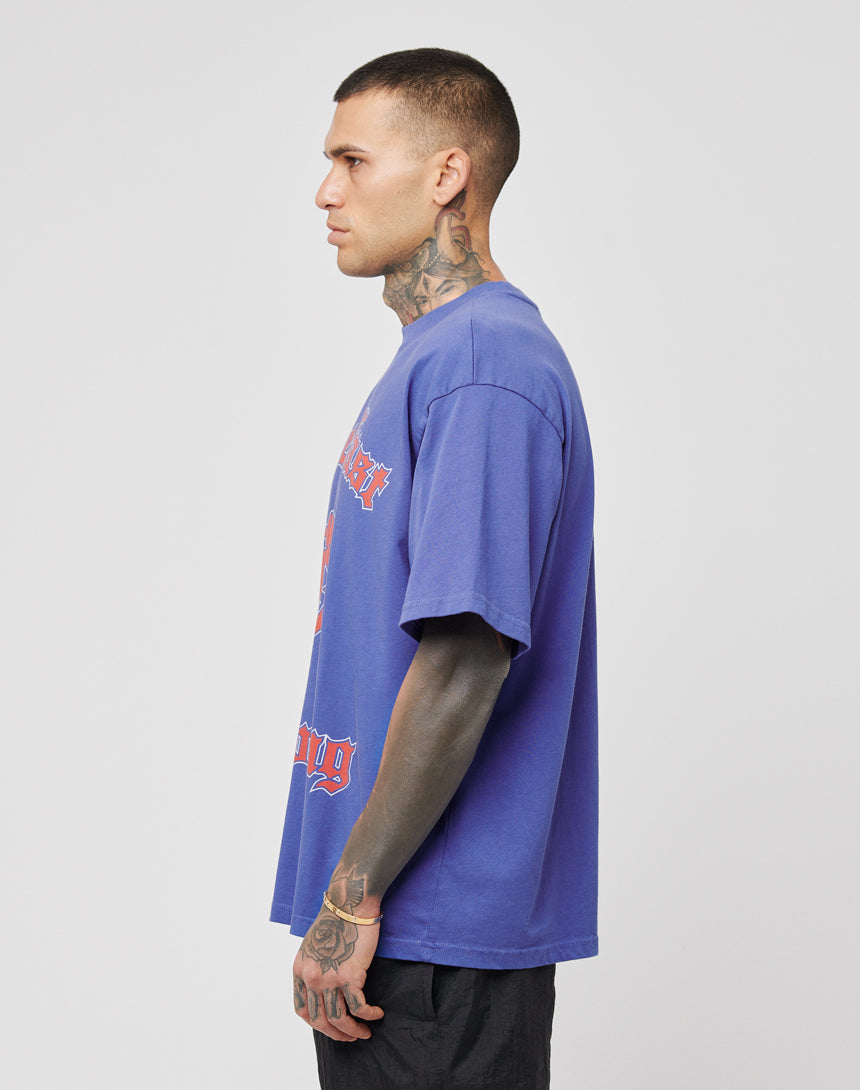 A tattooed individual stands in profile wearing the DY Tee by LIVE FAST DIE YOUNG and black pants against a plain light background.