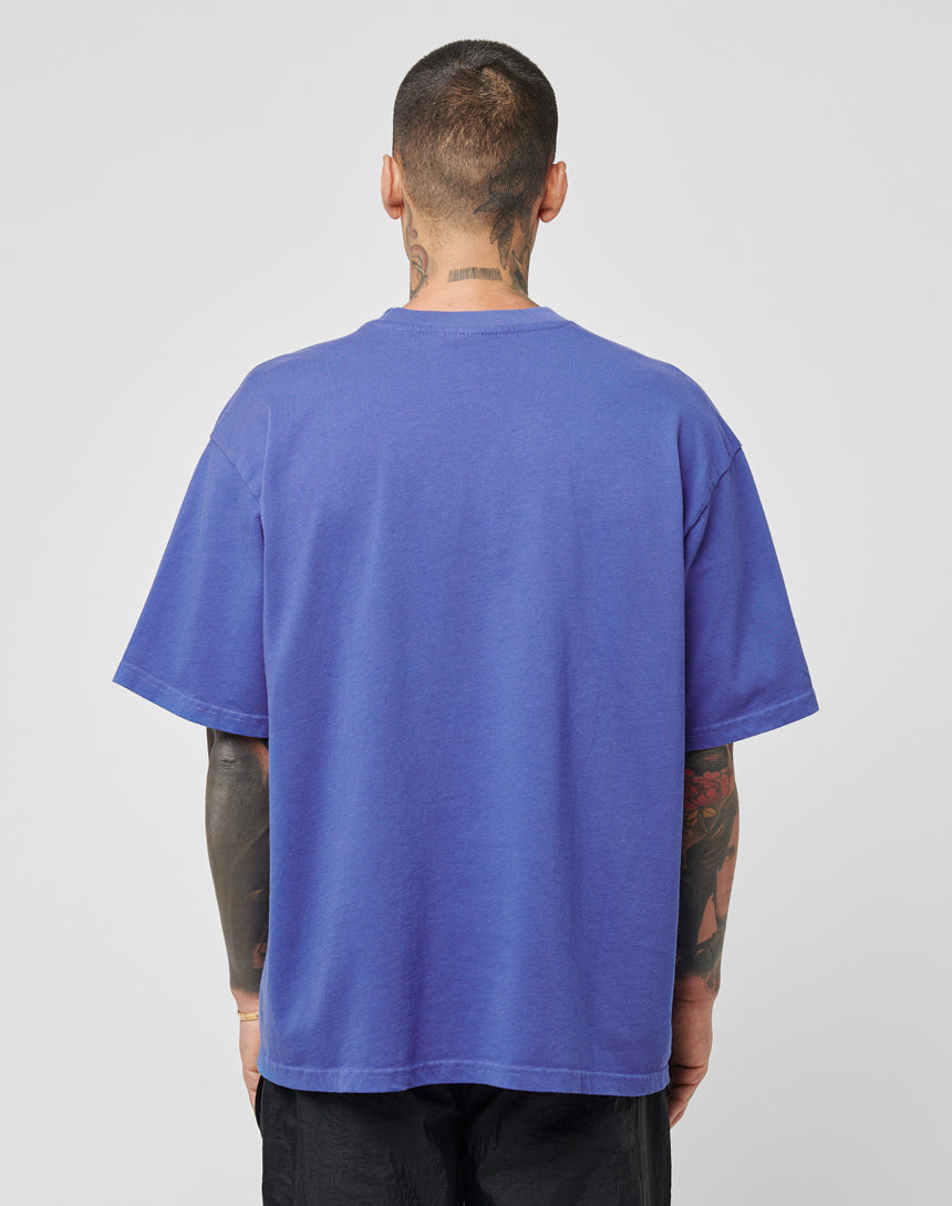 A person with short hair and visible tattoos faces away, wearing the LIVE FAST DIE YOUNG DY Tee in purple Vintage-Effekt (Regular Fit), paired with black pants against a plain light background.