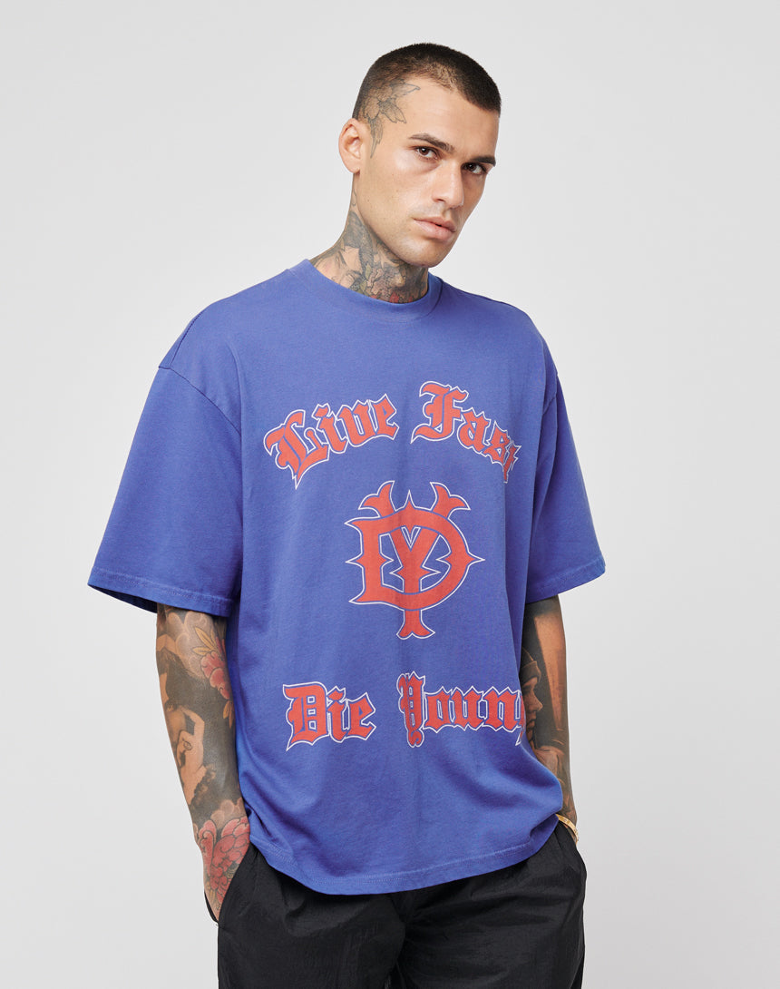 A tattooed man wears the DY Tee by LIVE FAST DIE YOUNG, a loose-fitting blue 100% Baumwolle T-shirt with red Gothic Live Fast Die Young print. With hands in pockets, he shows a cool vintage vibe and neutral expression.