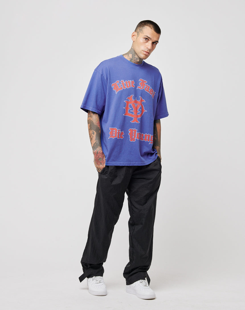 A young man with tattoos and short hair stands against a white background, wearing a LIVE FAST DIE YOUNG DY Tee—a loose blue graphic T-shirt with red lettering—paired with black pants and white sneakers. He has a neutral expression.