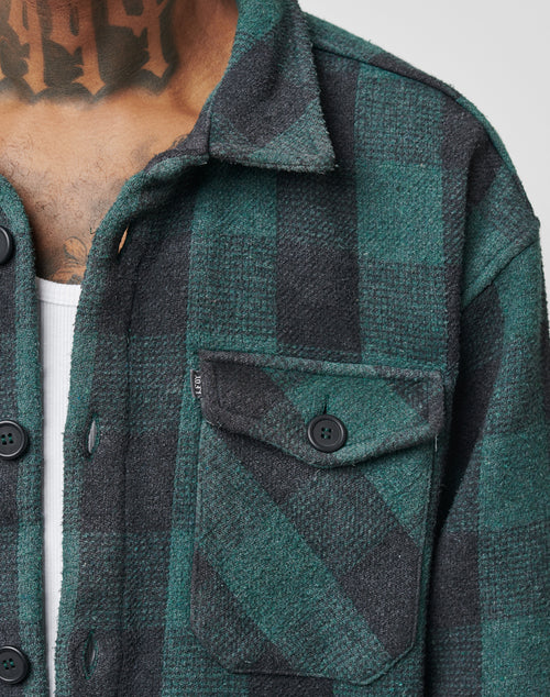 Close-up of a person in a LIVE FAST DIE YOUNG Heavy Flannel Shacket in green and black check, Regular Fit, over a white undershirt. Features black buttons, flap pocket, and part of a neck tattoo visible.