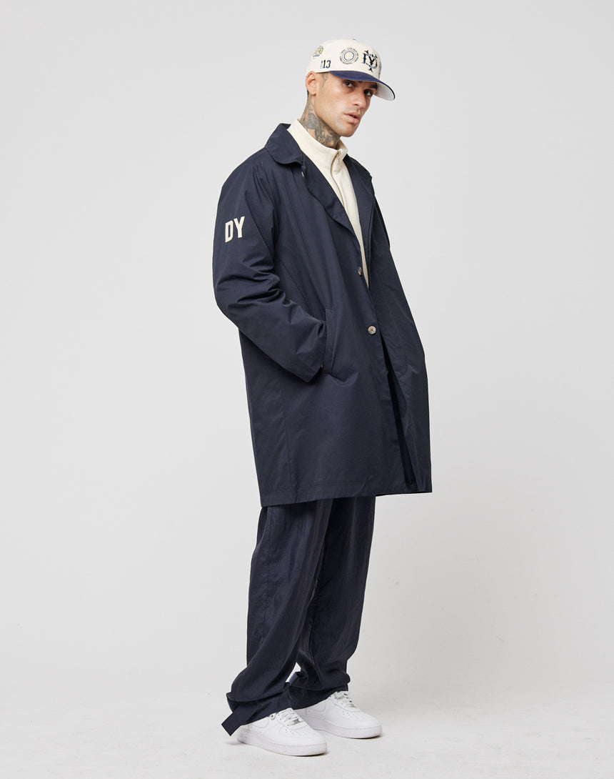 A person in a navy LFDY Trench Coat by LIVE FAST DIE YOUNG, with "DY" on the sleeve, a concealed button placket, navy pants, a white shirt, and a logo white cap stands against a plain white background looking downward.