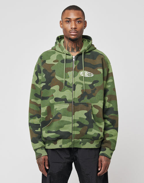 A man stands facing forward in a LIVE FAST DIE YOUNG LFDY Army Hooded Zip hoodie with green camouflage, Waffle Innenfutter, and NEEFAST chest logo, paired with black pants against a plain light background.