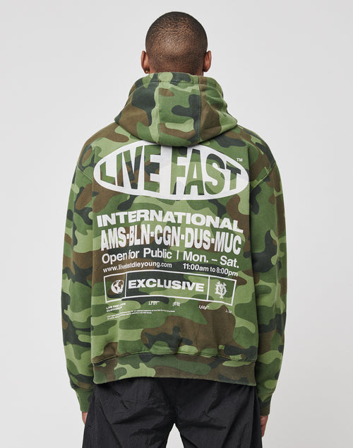 Someone stands against a plain light background wearing the LFDY Army Hooded Zip by LIVE FAST DIE YOUNG, a green camouflage hoodie with bold white LIVE FAST text on the back, Vintage-Effekt, and a list of international cities.