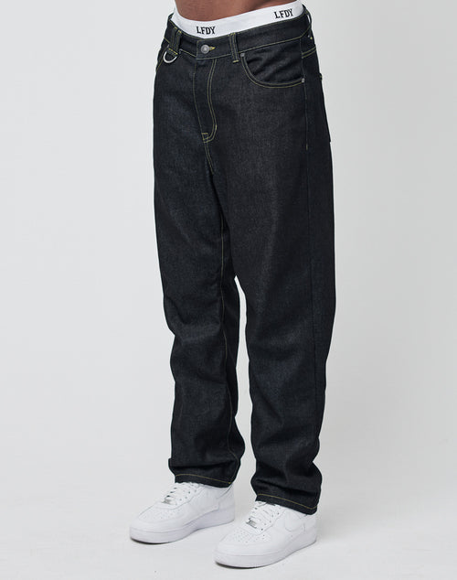 Someone wears loose-fitting Work Denim jeans by LIVE FAST DIE YOUNG, white sneakers, and shows the waistband of white LFDY underwear. The deep indigo denim stands out against a plain, light backdrop.