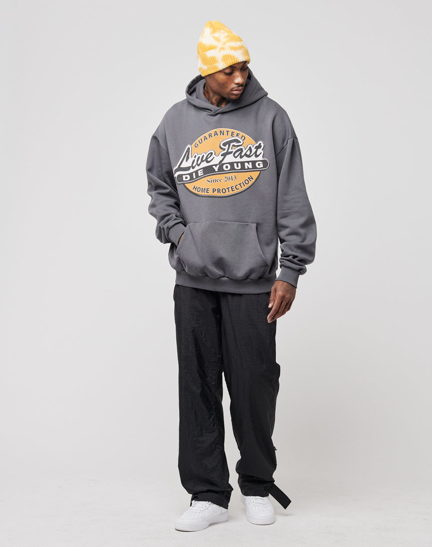 Wearing an oversize fit, dark gray Home Protection Hooded by LIVE FAST DIE YOUNG, black pants, a yellow-and-white beanie, and white sneakers, a person stands against a plain light background, gazing down to their left.