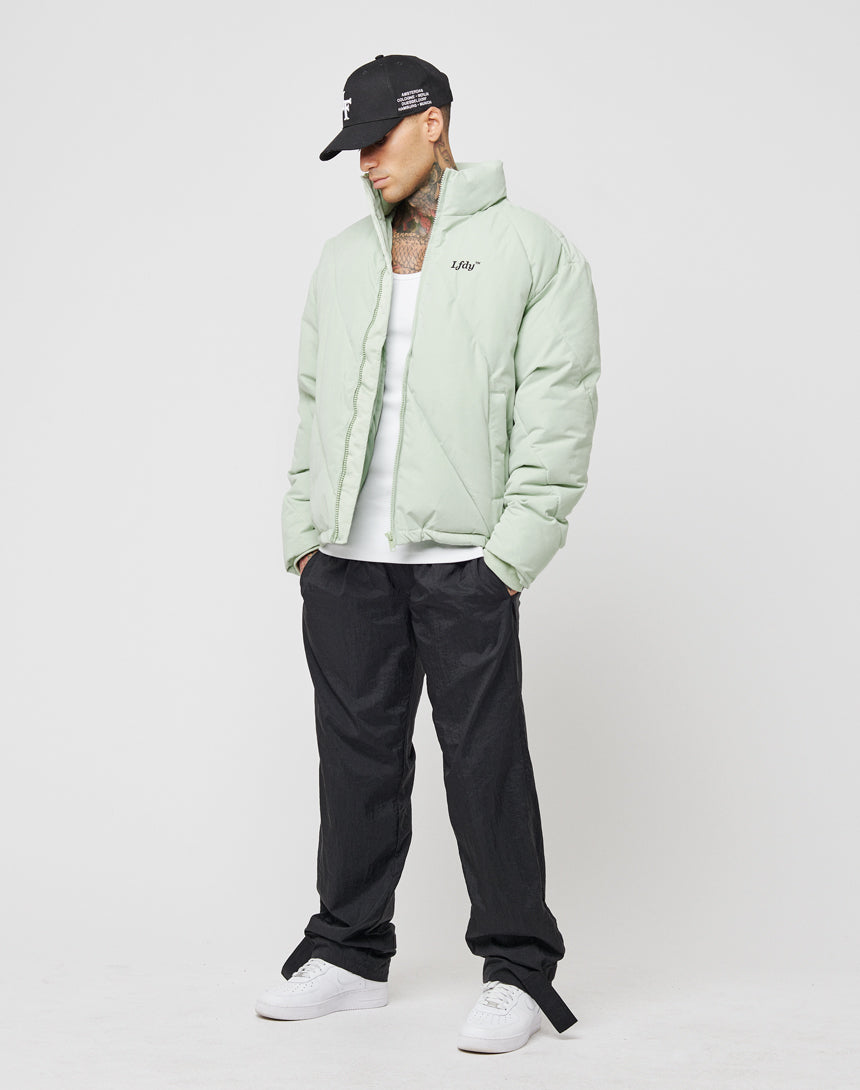A man in an LFDY light green Puffer Jacket with diagonal quilting, a white tank top, black pants, white sneakers, and a black cap stands against a plain white background looking down.