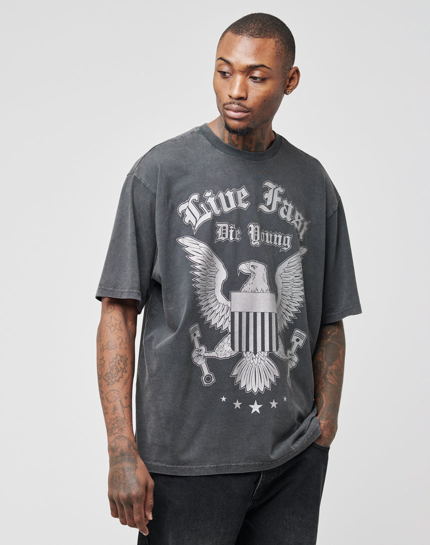 A man stands to the side with one hand in his pocket, wearing the Eagle Tee by LIVE FAST DIE YOUNG—a loose, faded black regular fit T-shirt with a bold eagle front print and slogan, made from 100% Baumwolle for all-day comfort.