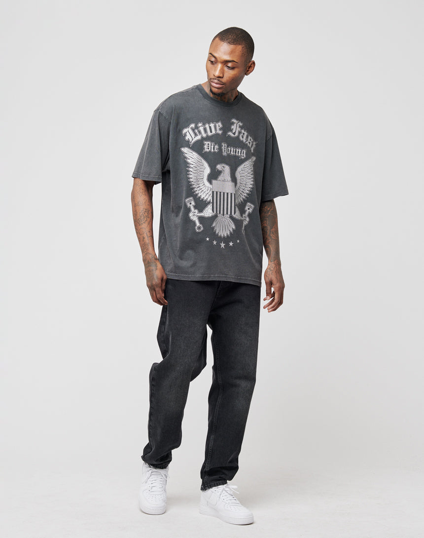 A man wears the Eagle Tee by LIVE FAST DIE YOUNG, a 100% Baumwolle, faded black regular fit T-shirt with a large eagle frontprint and Live Fast Die Young slogan. He pairs it with black jeans and white sneakers.