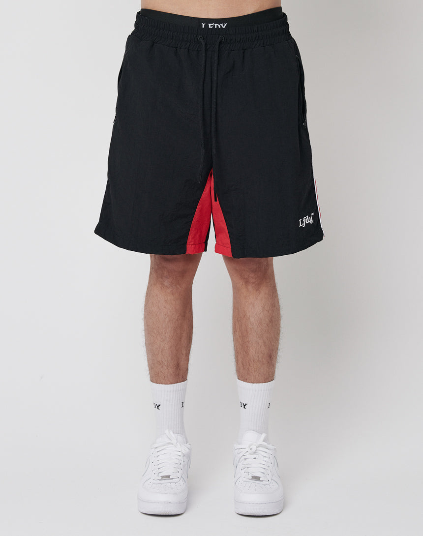 A person stands against a white background wearing LFDY Beach Shorts in black with red inner lining, an elastic waistband, and mesh inlay. The shorts feature a small white logo on the left leg, paired with white crew socks and sneakers.