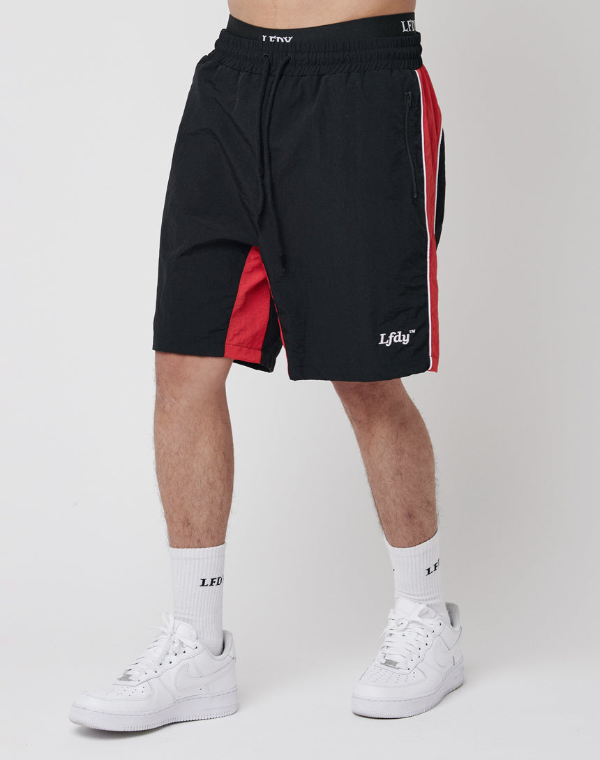 A person wearing LFDY Beach Shorts in black with red side panels and mesh inlay, featuring white LFDY text on the left leg, paired with white crew socks and sneakers, stands against a plain white background.