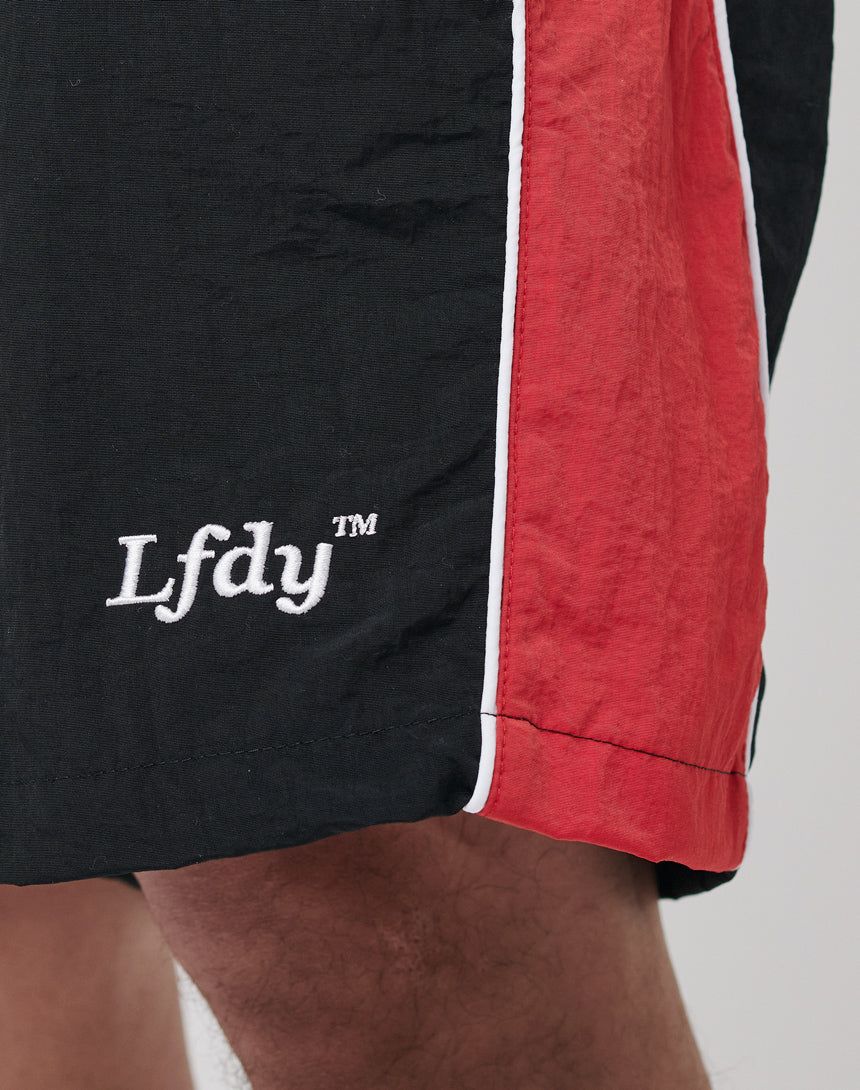 Close-up of a persons lower leg wearing LFDY Beach Shorts in black and red with a white stripe, featuring an elastic waistband and LFDY embroidered in white on the black section.
