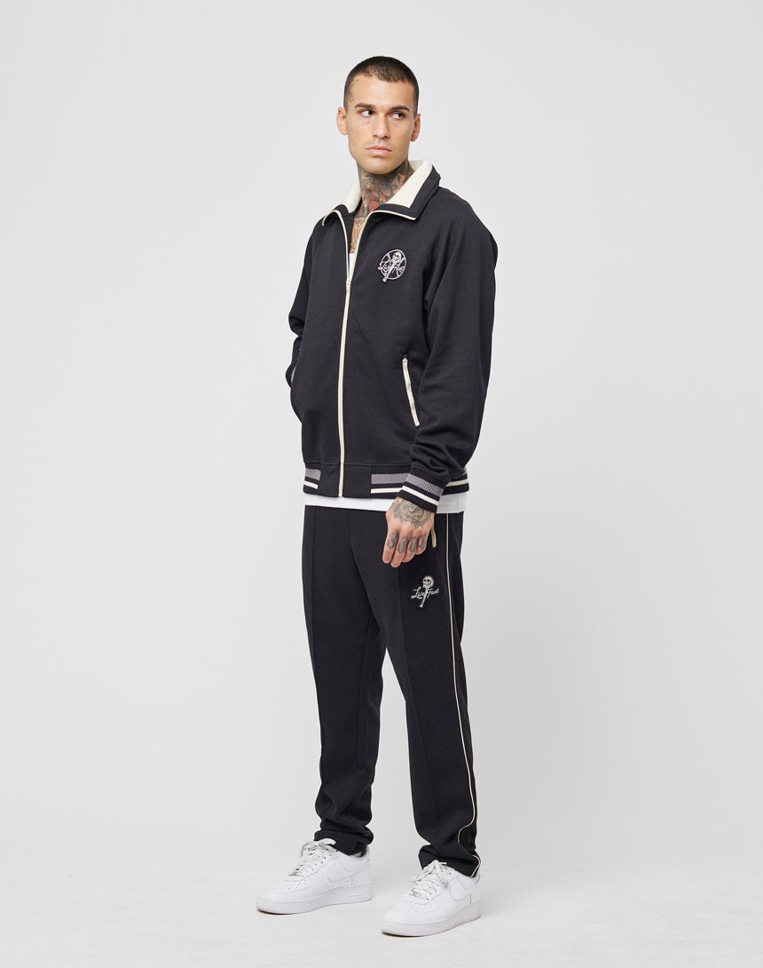 A man wearing a LIVE FAST DIE YOUNG Track Suit Jacket in regular fit black with white trim and white sneakers stands against a plain white background, looking slightly to his left with his hands in his pockets.