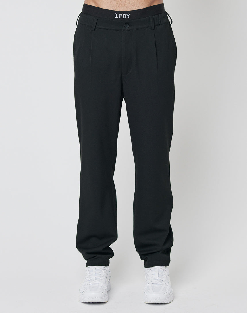 A person stands against a plain white background wearing LFDY Regular Casual Pants in black with pleats and an elastic waistband, paired with white sneakers.