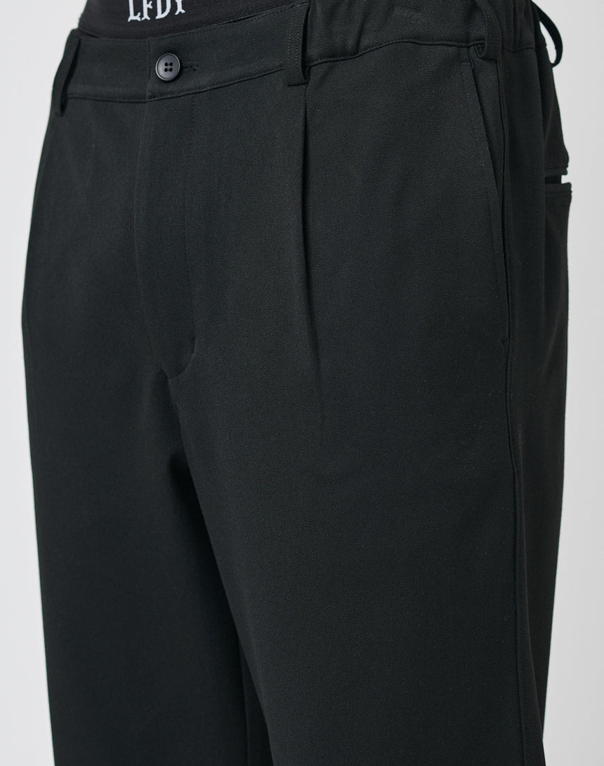 Close-up of a person wearing LFDY Regular Casual Pants in black, featuring a regular fit, pleated waistband, subtle side pockets, and an elastic waistband. A black waistband with white text is visible at the top.