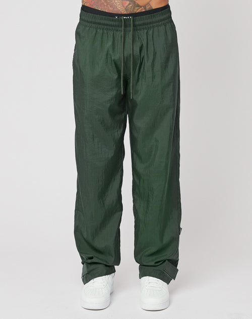 A person stands against a plain white background wearing LIVE FAST DIE YOUNG Nylon Jogger track pants in dark green with a wide elastic waistband, drawstring, and Flag Label detail, paired with white sneakers.
