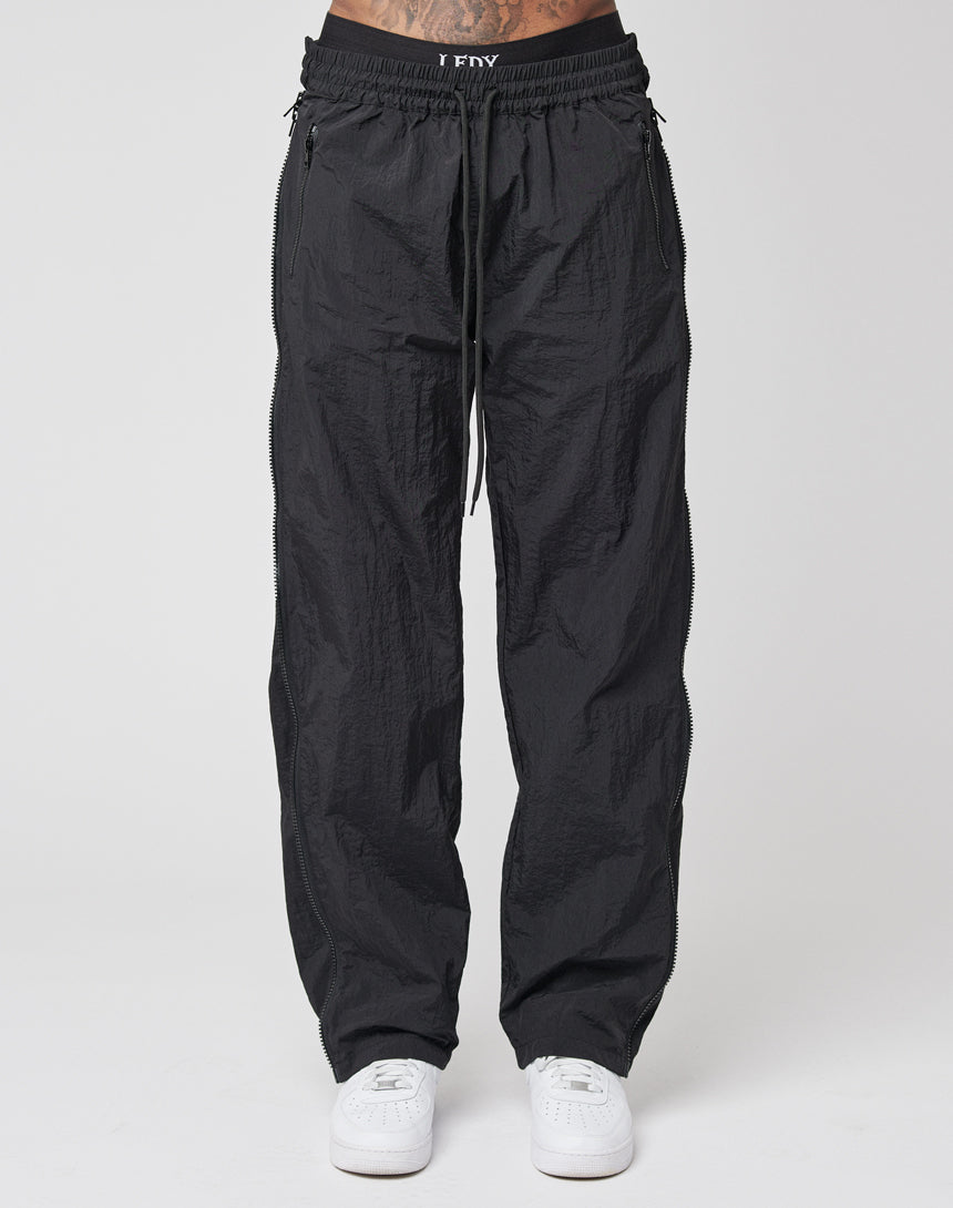 A person wearing LFDY Zip Tape Track Pants in black with a drawstring waist and white sneakers stands against a plain light background.