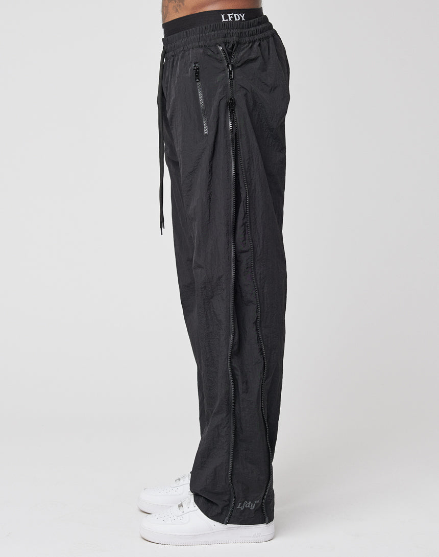 A person stands sideways against a plain white background wearing LFDY Zip Tape Track Pants, featuring branding on the waistband and ankle, paired with white sneakers.