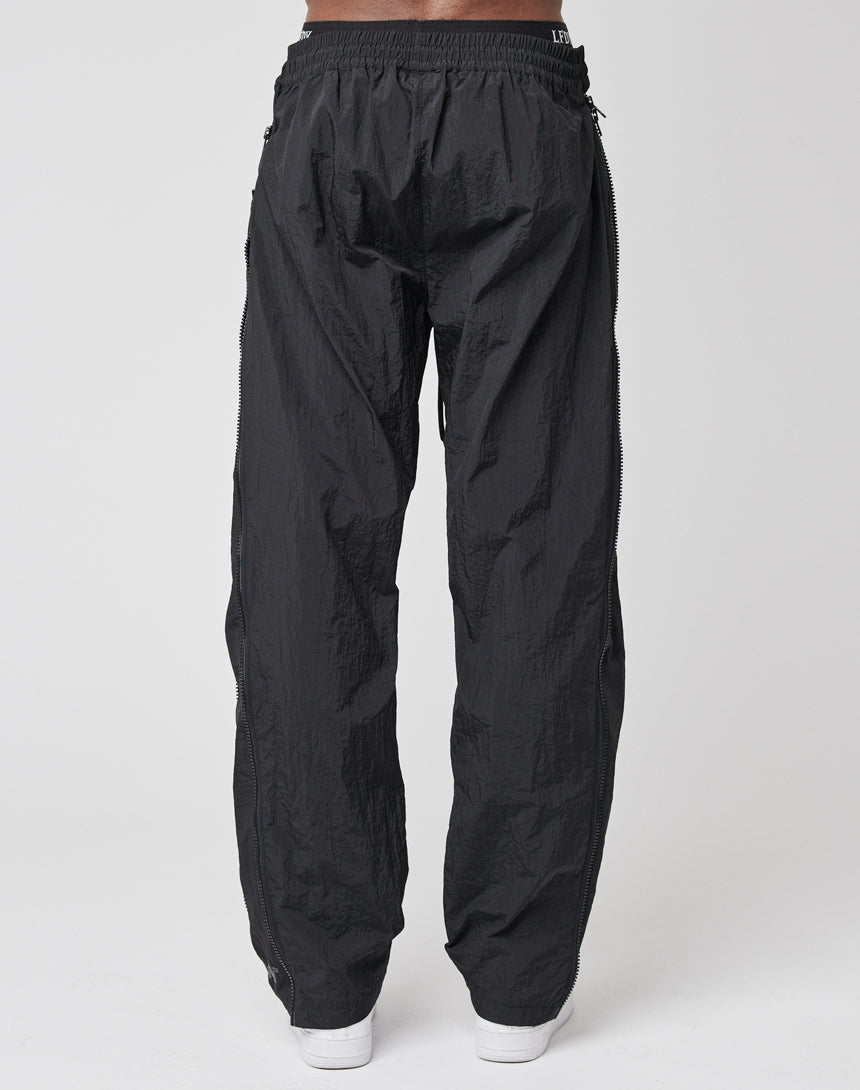 A person wearing LFDY Zip Tape Track Pants in black nylon with a crinkled texture and elastic waistband, viewed from the back. The regular fit pants are paired with white shoes against a plain white background.