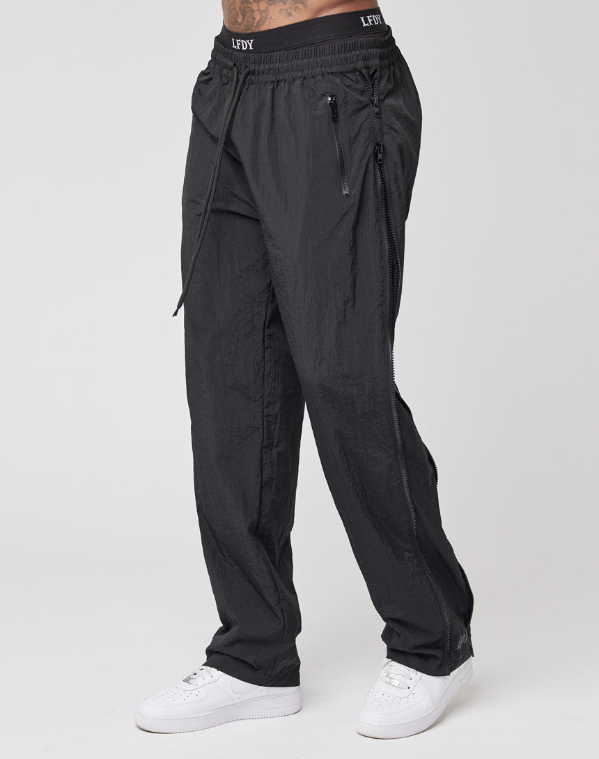 Person wearing LFDY Zip Tape Track Pants in regular fit black nylon with zipper pockets and LEFIX-branded waistband, paired with white sneakers, standing against a plain white background.